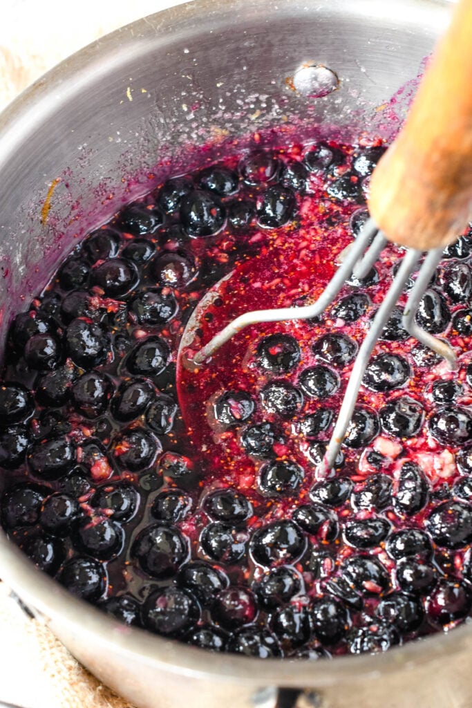 using a potato masher to squish the blueberries in the pan