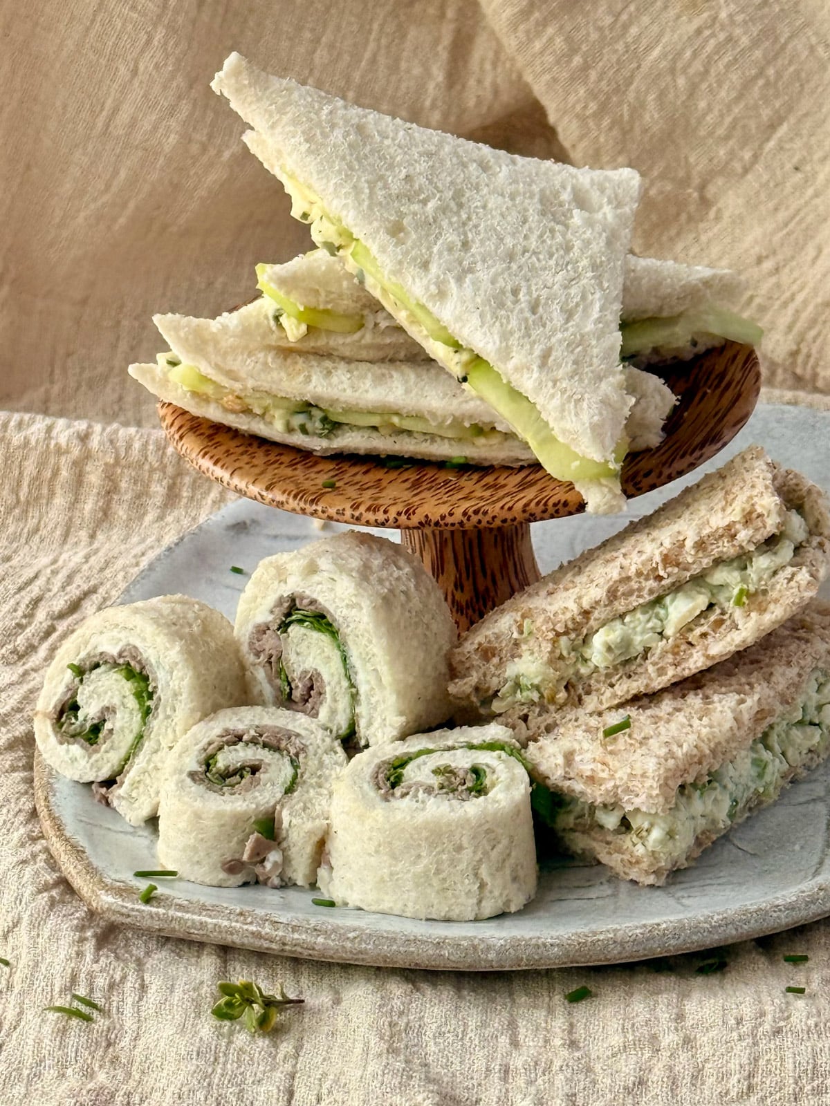 Assorted afternoon tea sandwiches and pinwheel sandwiches arranged on a plate and small wooden stand.