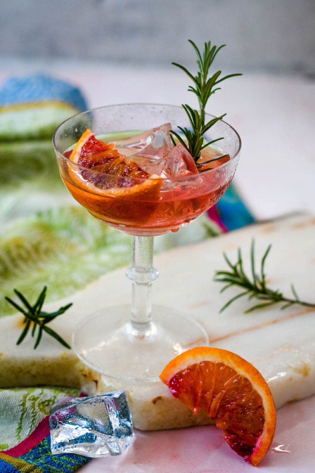 Blood orange gin cocktail with ice, blood orange slices, and a sprig of rosemary in a coupe glass.