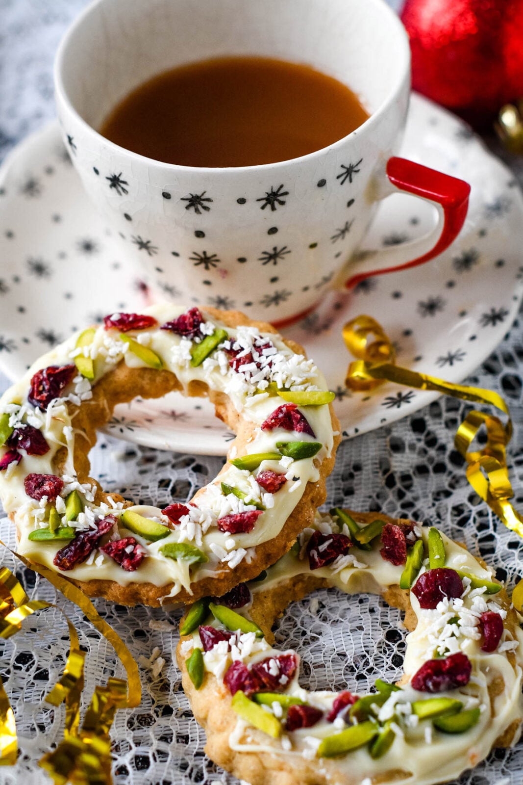 Christmas shortbread wreath cookies topped with white chocolate, pistachios, dried cranberries, and coconut beside a cup of tea.