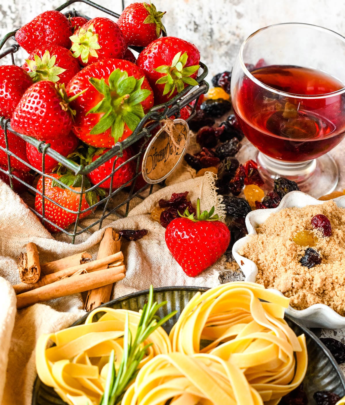 Strawberries, dried fruit, cinnamon sticks, brown sugar, pasta, herbs, and a glass of red wine arranged as cooking ingredients.