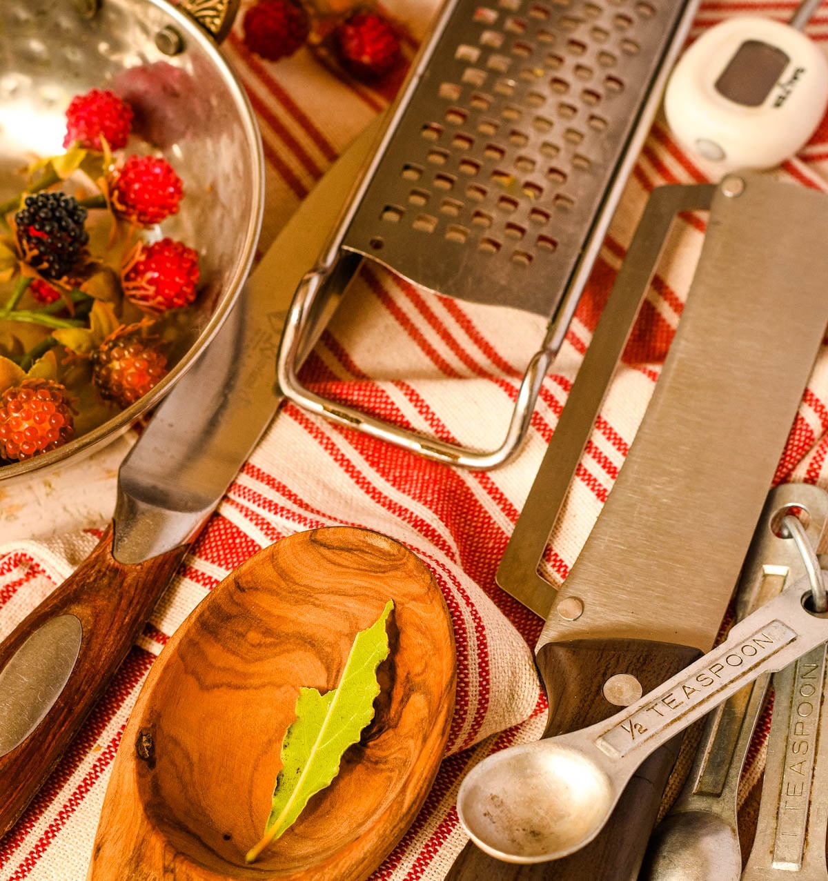 Kitchen tools including a grater, spatula, measuring spoons, timer, wooden utensil, and fresh berries on a striped cloth.