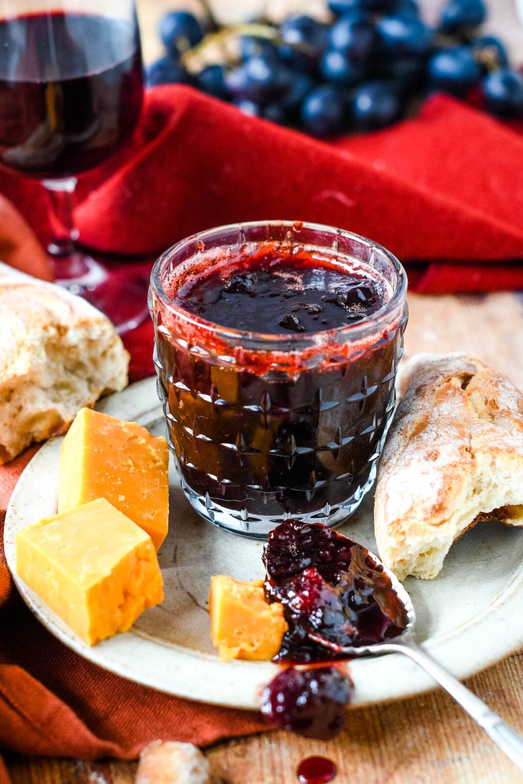 Mulled wine jam in a glass jar served with bread, cheese, grapes, and a glass of red wine.