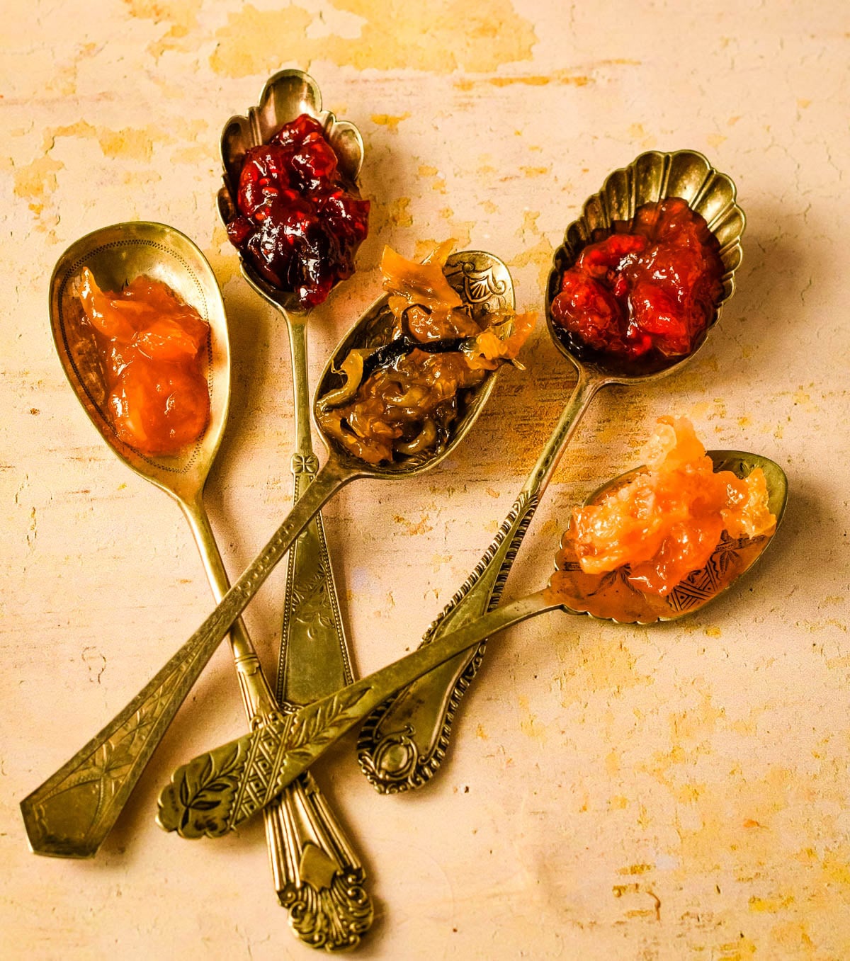 Vintage spoons filled with different homemade jams and preserves arranged on a rustic surface.