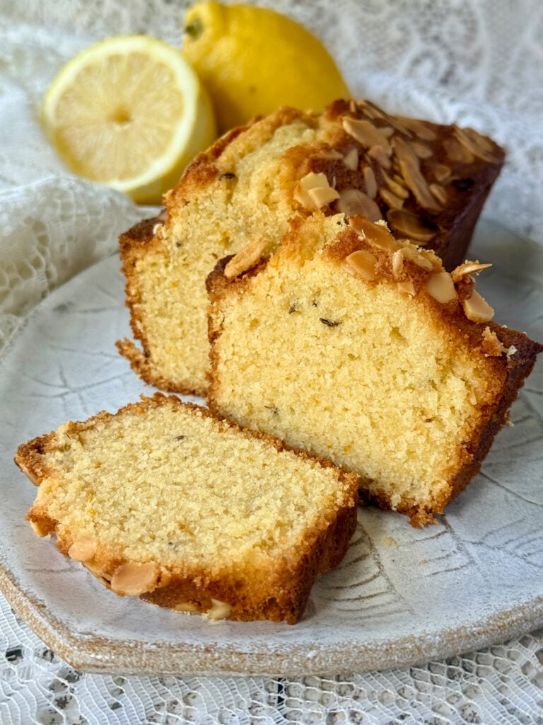 Slices of lemon loaf cake topped with flaked almonds and served with fresh lemons.